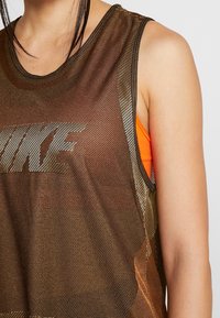 Camiseta sin mangas Nike en tela de malla marrón con un acabado brillante, con un logo blanco prominente y una camiseta interior naranja visible debajo.