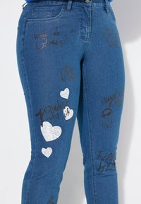 Blauwe denim jeans met verschillende gedrukte ontwerpen, waaronder zwarte tekst en witte harten, samen met een grafisch ontwerp van rolschaatsen.