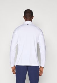 Weiße Langarm-Sportshirt mit hohem Kragen, glatter Textur und einem kleinen schwarzen Logo am Hals. Getragen mit dunkelfarbigen Hosen.