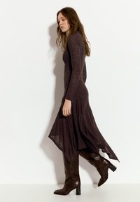 Lang, tætsiddende mørkebrun kjole lavet af struktureret stof, med lange ærmer og en asymmetrisk kant. Båret med knæhøje brune stiletter.