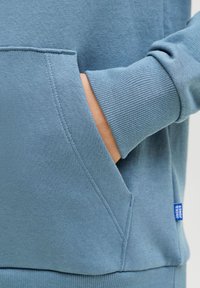 Χέρι μερικώς μέσα στην μπροστινή τσέπη ενός μπλε φούτερ με μακριά μανίκια, ελαστικές μανσέτες και μια μικρή μπλε ετικέτα Jack & Jones.