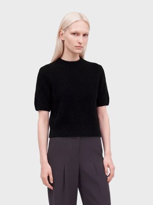 Personne aux longs cheveux blonds portant un pull noir à manches courtes et duveteux et un pantalon plissé sombre taille haute sur un fond clair uni.