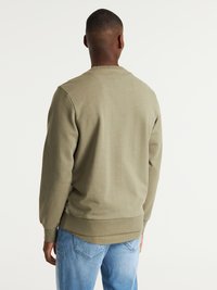 Sudadera verde oliva con puños y dobladillo acanalados, tejido texturizado y detalle de costura en la parte trasera, combinada con jeans de color azul claro.