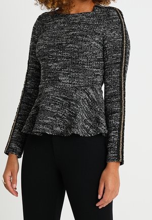 Femme portant un haut à manches longues texturé noir et blanc avec un basque et un détail chaîne le long des manches, associé à un pantalon noir.