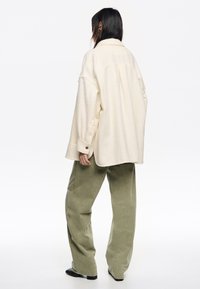 Camisa de forro polar de color crema y corte oversize, con cuello y puños abotonados, combinada con pantalones de pana sueltos en un tono verde claro.