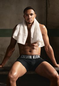 Melni Calvin Klein boksa šorti ar baltu jostu. Modelis sēž uz sola ar dvieli, kas pārsvērts pār pleciem.