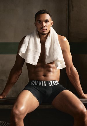 Zwarte Calvin Klein boxershorts met een witte tailleband. Het model heeft een handdoek over zijn schouders gedrapeerd en zit op een bankje.