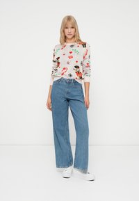 Gestickter Pullover mit einem hellbeigen Hintergrund, der bunte, festliche Muster zeigt. Kombiniert mit weit geschnittenen blauen Jeans und weißen Sneakers.
