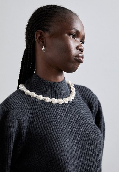Collier de perles avec des perles rondes et embellies ; porté sur un pull en maille côtelée foncé, avec un col montant et un tissu texturé.