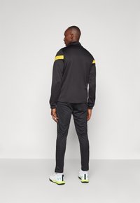 Black athletic tracksuit with yellow shoulder accents, featuring a high collar, zipped front, and tapered pants. White shoes with green details.