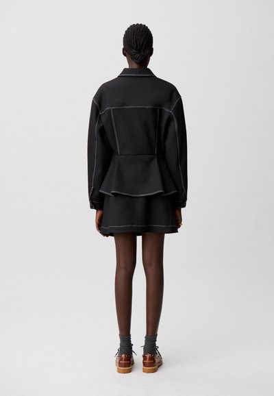 Completo con giacca nera e gonna con cuciture bianche a contrasto, vita a peplo, tessuto liscio e scarpe marroni alla caviglia.