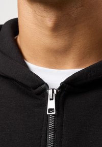 Zwarte hoodie met ritssluiting in zilver en een wit binnenlaagje zichtbaar bij de halslijn. Heeft een zachte textuur en een nauwsluitend ontwerp.