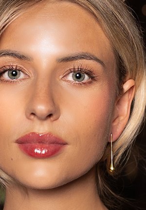 Gouden druppeloorbel bij een vrouw met een egale huid, lichtblond haar en glanzende rode lippen. Close-up benadrukt de gelaatskenmerken.