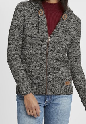 Mujer con sudadera con capucha de punto gris y negra con cremallera sobre una camiseta burdeos acanalada y vaqueros azules, de pie con la mano izquierda relajada.