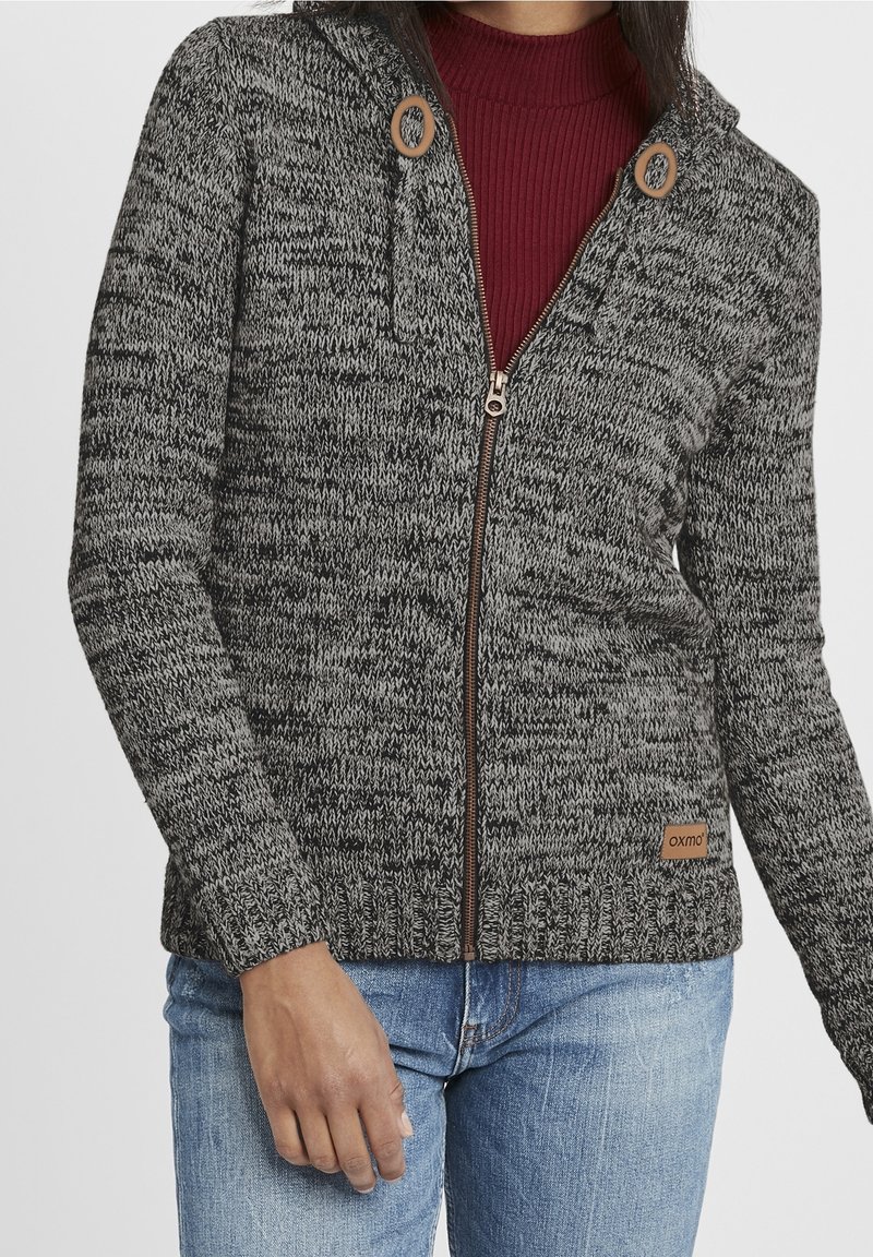 Femme portant un sweat à capuche zippé en tricot gris et noir sur un haut côtelé bordeaux et un jean bleu, debout, main gauche détendue.