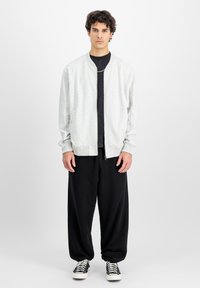 Svetlo siva bomber jakna z okroglim ovratnikom in rebrastimi manšetami, nosena čez temno srajco, v kombinaciji z ohlapnimi črnimi hlačami in supergami.