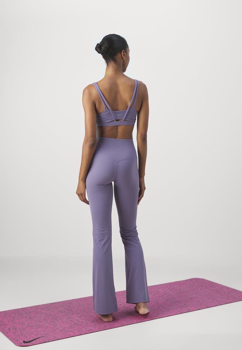 Sujeción deportiva morada con tirantes entrecruzados, combinada con mallas moradas acampanadas a juego, sobre una esterilla de yoga de color rosa con manchas.