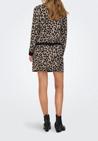 Femme portant une robe beige et noire à imprimé léopard avec des manches longues et des bottines noires, debout de dos sur un fond uni.