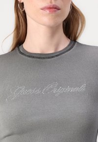 Grå ribbet topp med rund halslinning som har sølv rhinestone-tekst "Paris Original" over brystet. Glatt tekstur og tettsittende design.