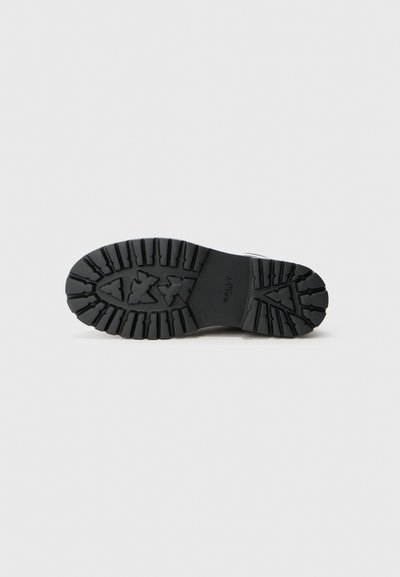 Semelle de chaussure en caoutchouc noir avec un motif de relief profond et un logo "Bally" au centre, vue du dessous sur un fond uni.