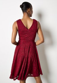 Un vestido de encaje burdeos con escote en V, diseño sin mangas y falda acampanada. La tela presenta un patrón floral y una cremallera oculta en la parte posterior.