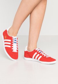 Röda mockaskor med vit tre-stripes-design och vit gummisula. Har vita snörningar och blå detaljer på tungan.