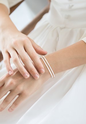 Brazalete blanco con detalles en oro, con un diseño intrincado de cadenas, llevado en una muñeca ligeramente bronceada. Acabado suave y brillante.