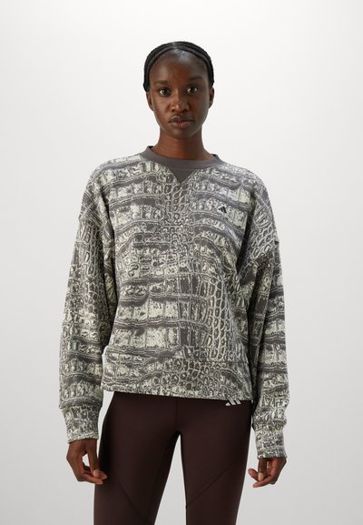 Ung kvinne med flettet hår iført en grå og hvit mønstret langermet skjorte og mørke leggings, stående mot en enkel hvit bakgrunn.