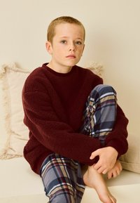 Maranfleece villapaita ja ruudulliset pyjamahousut sinisessä, harmaassa ja beigeessä. Lapsi istuu vaalealla sohvallä, jossa on fringevilttiä, paljain jaloin.