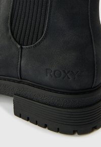 Ghete negre de gleznă din material de piele întoarsă texturată, cu panouri elastice striate pe laterale și talpă robustă din cauciuc. Prezintă logo-ul „ROXY” în relief.