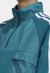 Tyrkysová poloviční zipová bunda s vysokým límcem, se bočními pruhy v bílé barvě, texturovaným materiálem a přední kapsou.