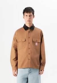 Joven vestido con una camisa marrón de botones y cuello negro, con dos bolsillos en el pecho y jeans de color azul claro, de pie frente a un fondo blanco.