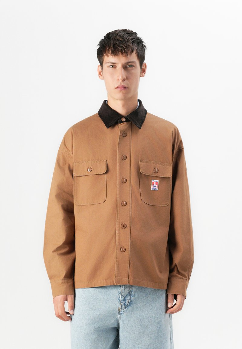 Young man wearing brown button-up shirt with black collar, two chest pockets, and light blue jeans, standing against a white background.