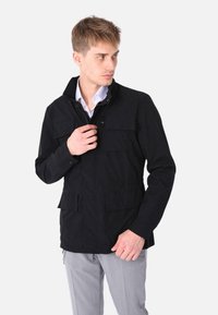 Giacca nera con colletto, multiple tasche frontali e chiusura con zip; indossata sopra una camicia azzurro chiaro, mostrata su un modello.