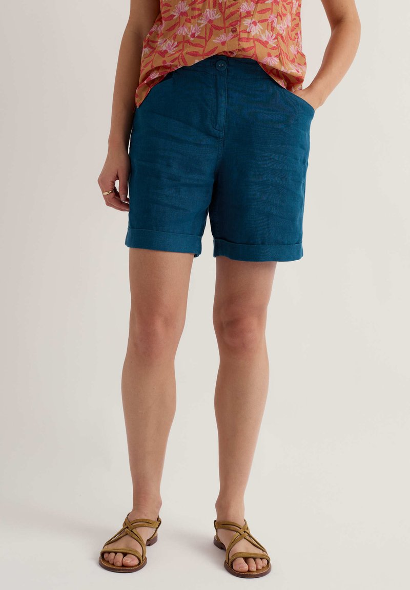 Frau trägt kurz geschnittene Shorts mit umgekrempeltem Saum in Türkis, ein oranges, geblümtes Oberteil und braune flache Sandalen mit Riemchen, steht mit einer Hand in der Hosentasche.