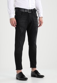 Černé džíny z denimu s proříznutými detaily, zúžený střih, design se pěti kapsami, nošeny s bílou košilí a černými koženými botami.