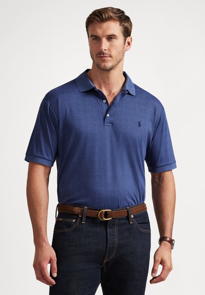 Άνδρας που φοράει μπλε πουκάμισο πόλο με καρό σχέδιο, καφέ ζώνη, σκούρο τζιν και καφέ ρολόι χειρός, στέκεται μπροστά σε ένα απλό φόντο.