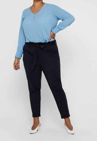 Person wearing a light blue long-sleeve blouse, black high-waisted tapered pants with a tie belt, and white low-heeled shoes, standing.