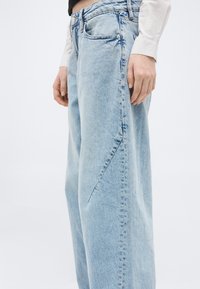 Φαρδιά τζιν παντελόνια ανοιχτού γαλάζιου χρώματος που φοριούνται από ένα άτομο με λευκό πουκάμισο μακρυμάνικο, μπροστά από τη μέση έως τα πόδια.