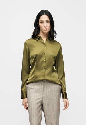 Oliivinvihreä satiinipusero, jossa pitkät hihat, kaulus ja napitettu edusta. Yhdistetty vaaleanharmaisiin raidallisiin housuihin, sileä pinta.