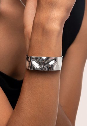 Silberne Armreifen mit einer glatten, reflektierenden Oberfläche und einem breiten, minimalistischen Design. Sitzt eng am Handgelenk.