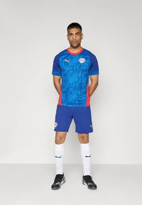 Modra in rdeča nogometna oprema znamke Puma z modrim zgornjim delom z teksturo in logotipom, modrimi hlačami, belimi nogavicami ter črnimi nogometnimi čevlji.