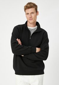 Svart zip-up sweatshirt med hög krage, ribbade manschetter och nederdel. Har ett mjukt, strukturerat tyg och bärs över en randig skjorta.