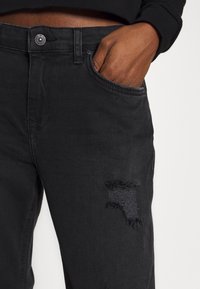 Jeans de mezclilla negra con un parche desgastado en el muslo. Presentan un diseño clásico de cinco bolsillos y un cierre de un solo botón.