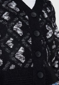 Mustat neulottu villatakki suurilla mustilla napilla ja valkoisia sekä harmaita sahalaitakuvioita kankaan päällä.