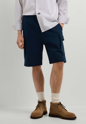 Man draagt marineblauwe cargoshorts, witte sokken, bruine laarzen en een licht gestreept shirt met lange mouwen, met één hand in de zak.