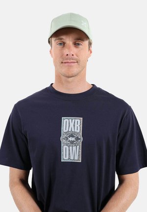 Homme portant une casquette vert clair et un t-shirt bleu marine avec un motif rectangulaire blanc et bleu affichant "OXB OW" sur la poitrine, devant un fond uni.