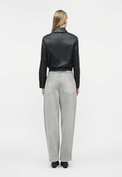 Γυναίκα με την πλάτη στραμμένη, φοράει μαύρο cropped δερμάτινο μπουφάν και ανοιχτό γκρι ψηλόμεσο παντελόνι με πλατιά μπατζάκια, μαζί με μαύρα μποτάκια αστραγάλου.