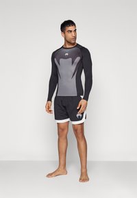 Langermet grå og svart rash guard med en tettsittende design, sammen med svarte shorts med hvit kant og logo på benet.
