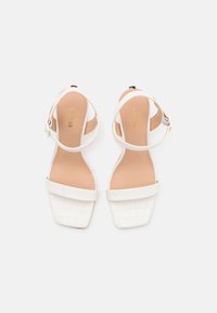 Sandali bianchi con tacco, in pelle di coccodrillo, punta quadrata e cinturini alla caviglia. Soletta beige con logo impresso. Design morbido e strutturato.